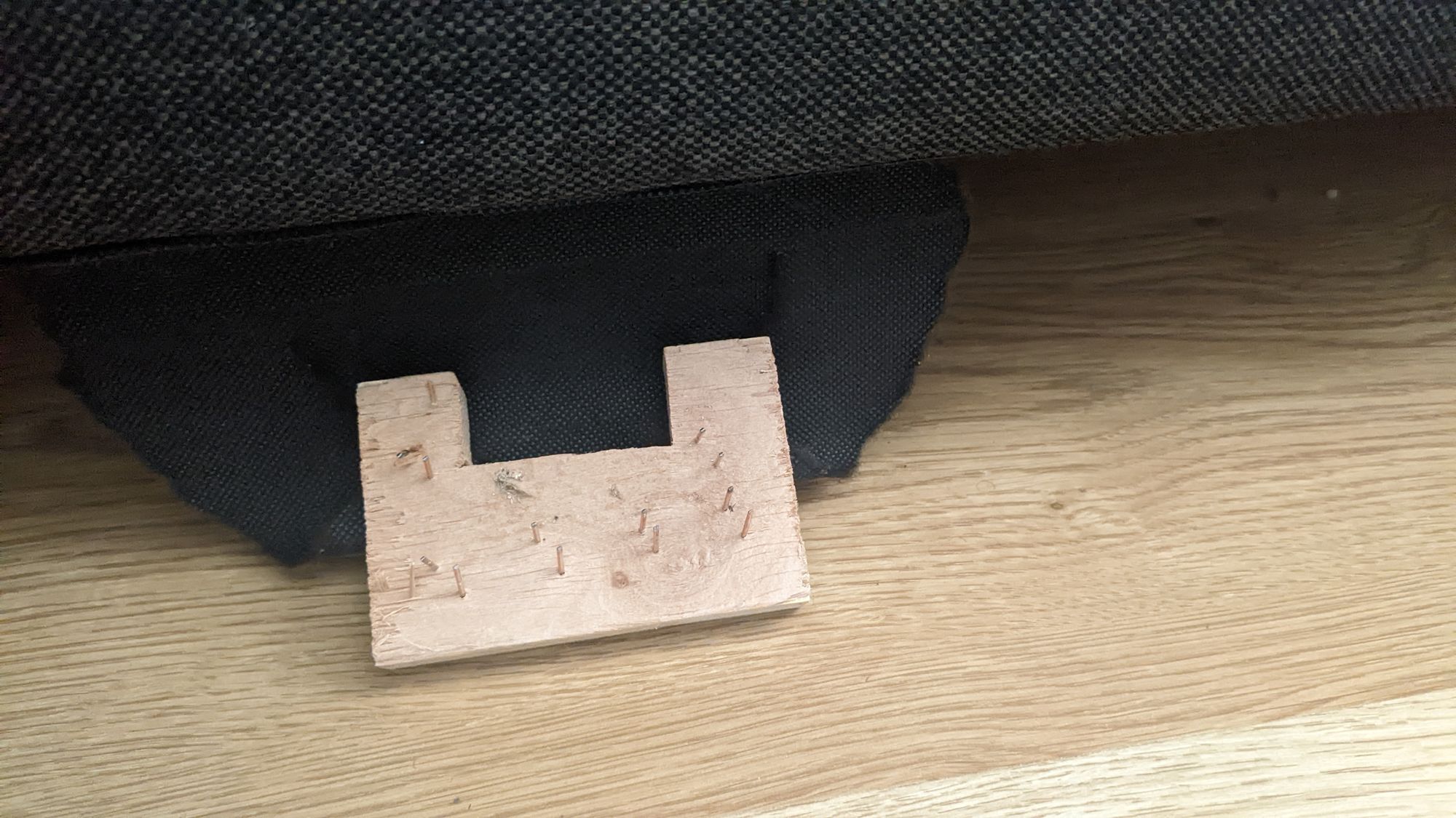 Picture of a wood block connected to some cloth with nails sticking out of it, underneath a sofa