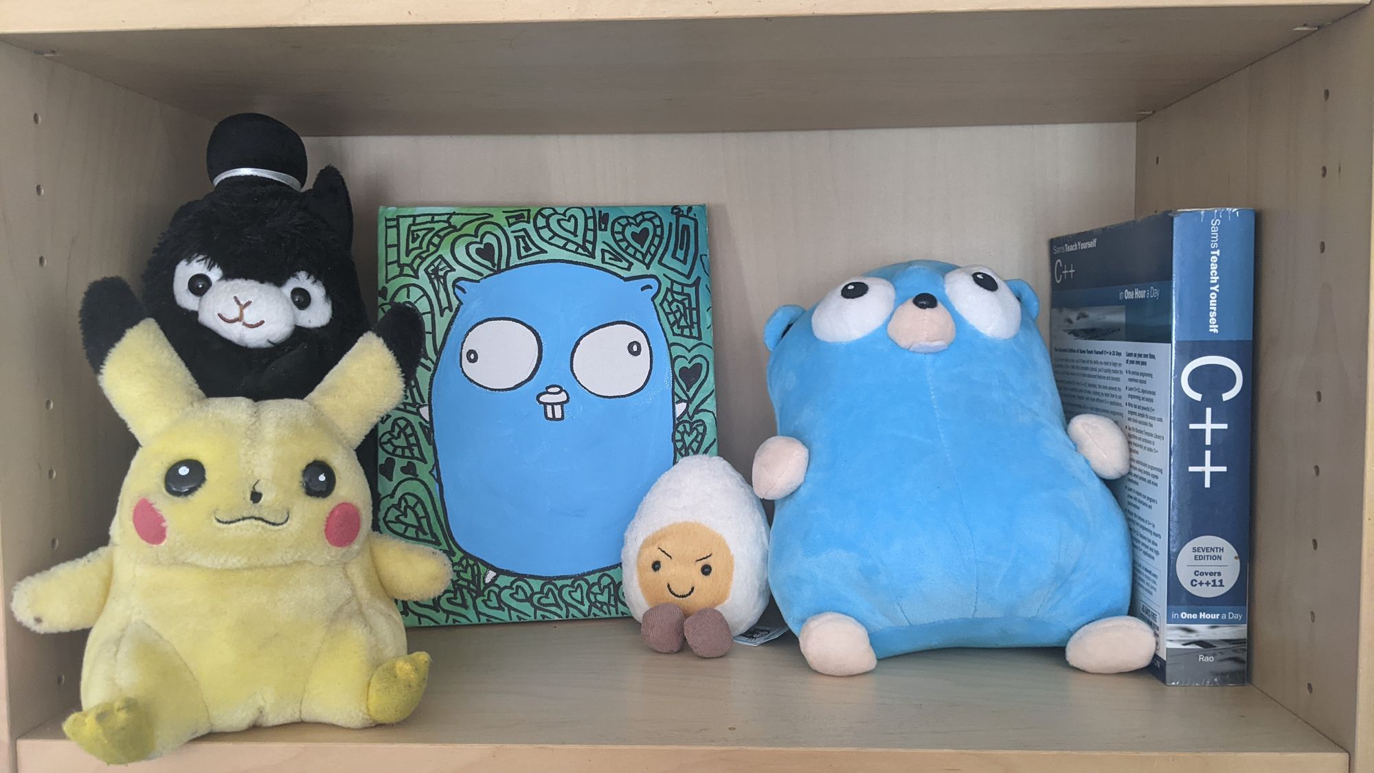 A collection of cuddly toys, including a pikachu, llama with a hat, an egg and a big blue gopher. There is also a picture of the blue gopher and a c++ book